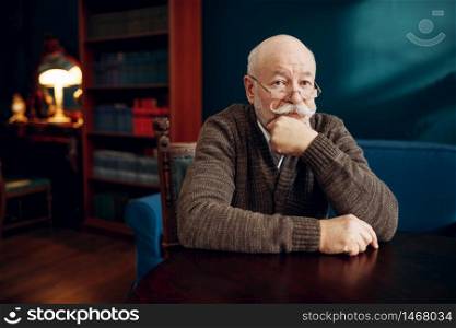 Serious elderly man sitting at the table in home office. Bearded mature senior in living room, old age businessman. Elderly man sitting at the table in home office