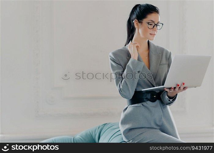 Serious elegant female administrator works from home, reading or watching a webinar on laptop, dressed in stylish business clothes, leaning on a sofa.