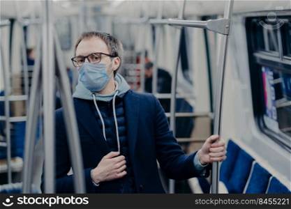 Serious European man being aware of his health, protects from catching serious disease in public transport, wears medical mask on face, travels in urban train, afraids of coronavirus epidemy