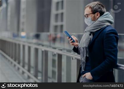 Serious European man looks in smartphone, reads news online, sends text messages, being protected from virus, wears medical mask, cares about public coronavirus protection. Virus outbreak, Covid-19