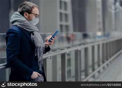 Serious European man looks in smartphone, reads news online, sends text messages, being protected from virus, wears medical mask, cares about public coronavirus protection. Virus outbreak, Covid-19