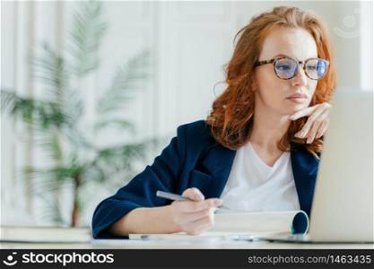 Serious experienced female coach makes records in notepad, focused at screen of laptop computer, has long red hair, wears transparent glasses, watches webinar online. Business and job concept