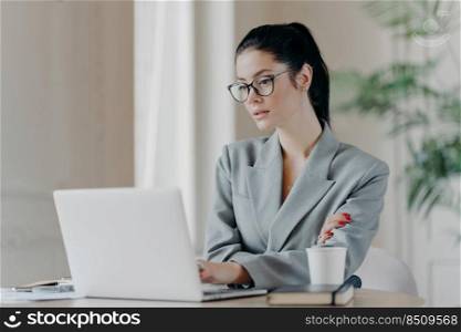 Serious female entrepreneur in eyeglasses, has dark hair combed in pony tail, works on business project, concentrated at laptop computer wears grey formal costume uses information from web resources