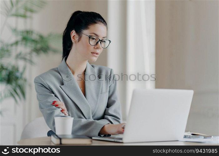 Serious female entrepreneur in glasses, ponytail, and grey formal attire, focused on laptop, utilizing web resources for business project.