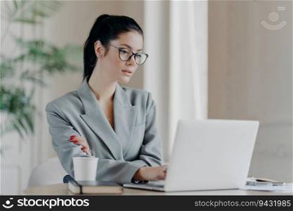 Serious female entrepreneur in glasses, ponytail, and grey formal attire, focused on laptop, utilizing web resources for business project.