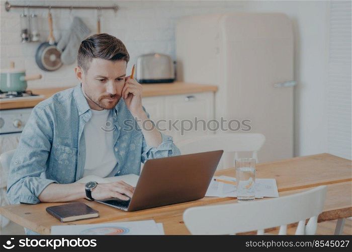 Serious focused determined young bearded businessman or freelancer in casual clothes sitting at kitchen table and typing on laptop, working on project online, researching or writing email to client. Serious focused bearded businessman in casual clothes working on laptop while sitting at kitchen table