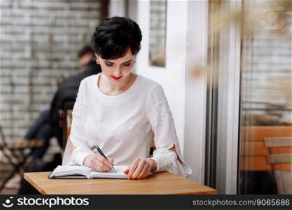 Serious focused smart businesswoman is writing notes on notepad, making agenda on personal organizer. Female in business suit sitting at table at cafe on terrace, work and event planning. Serious focused smart businesswoman is writing notes on notepad, making agenda on personal organizer. Female in business suit sitting at table at cafe on terrace, work and event planning.