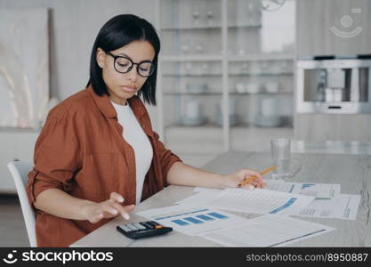 Serious girl, accountant is examining reports. Young european woman in glasses is working with data charts from home. Manager or business assistant is sitting at the desk and doing business.. Accountant examining reports. Business assistant in glasses is working with data charts from home.