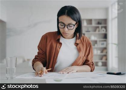 Serious girl, businesswoman is examining reports. Young european woman in glasses and casual wear is working remote from home. Manager or business assistant is sitting at the desk and doing paperwork.. Serious girl, businesswoman is examining reports. Young european woman is working remote from home.