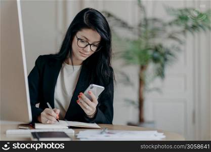 Serious good looking adult woman with dark hair, concentrated into mobile phone, searches necessary information in internet, writes down notes in notepad, sits at desktop, surrounded by papers