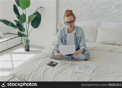 Serious good looking redhead European woman calculates expenses manages household budget looks attentively at paper documents wears denim casual clothes sits on bed against cozy home interior. Good looking redhead European woman calculates expenses manages household budget