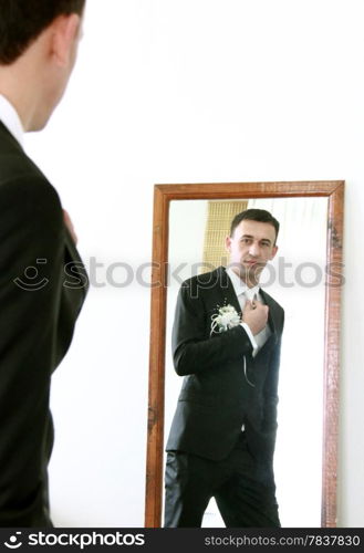 Serious handsome bridegroom looking in mirror straightening his tie