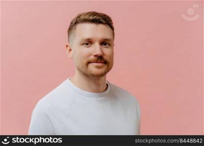 Serious handsome European man with stubble looks directly at camera dressed in casual white t shirt has calm face expression poses against pink background. Bearded guy ready for having walk.