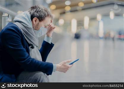 Serious infected man wears protective mask, cares about health during coronavirus outbreak, uses mobile phone, reads information online about covid-19, checks news, poses indoor. Information panic