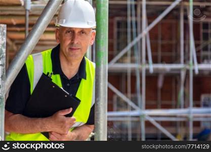 Serious looking male builder foreman, worker, contractor or architect on construction or building site holding black clipboard wearing white hard hat