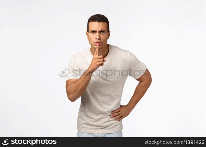 Serious-looking strong young muscular man demand silence, shushing with index finger near lips, frowning telling keep quiet, have rules be silent, hush at camera, white background.. Serious-looking strong young muscular man demand silence, shushing with index finger near lips, frowning telling keep quiet, have rules be silent, hush at camera, white background