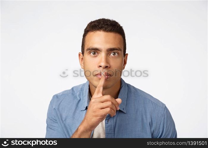 Serious-looking young hispanic man bending towards camera, shushing, make shush or shh sound as press finger to lips, look assertive, ask stay quiet, demand silence, white background.. Serious-looking young hispanic man bending towards camera, shushing, make shush or shh sound as press finger to lips, look assertive, ask stay quiet, demand silence, white background