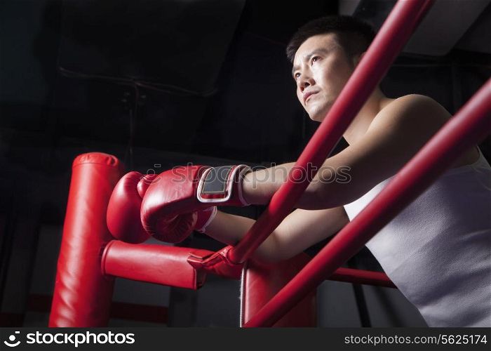 Serious male boxer resting his elbows on the ring side, looking away, low angle view