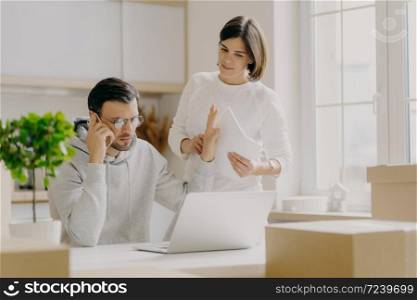 Serious male calls via smartphone, makes refusal gesture, wears spectacles and sweatshirt, asks wife to wait, tries to decide financial problems, pose in kitchen with big windows surrounded with boxes