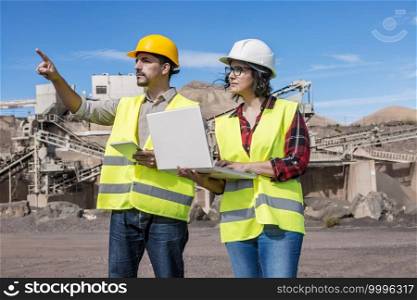 Serious male engineer with tablet pointing away and asking opinion of female colleague with laptop while developing construction plan for industrial project. Construction engineers with laptop and tablet inspecting industrial facility