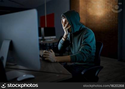 Serious male internet hacker in hood works on computer, front view. Illegal web programmer at workplace, criminal occupation. Data hacking, cyber security. Serious male hacker in hood works on computer