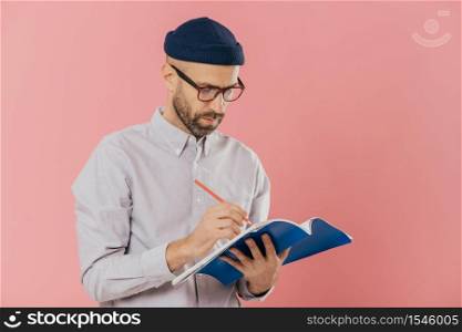 Serious male writer writes essay in notepad, feels inspired, wears glasses and formal clothes, preapares creative ideas for new book, models against pink studio wall. Bearded man with organizer