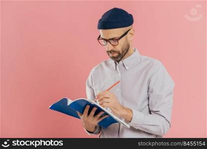 Serious male writer writes essay in notepad, feels inspired, wears glasses and formal clothes, preapares creative ideas for new book, models against pink studio wall. Bearded man with organizer