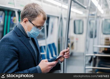 Serious man commutes to work in underground wears virus protective mask being always in touch with modern cellular poses in empty subway or metro. Distance concept. Public transport during coronavirus