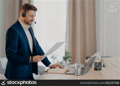 Serious man excutive worker gives presentation of business plan holds paper document stands near desktop dressed formally uses modern laptop and headset for online conference. Social distance working