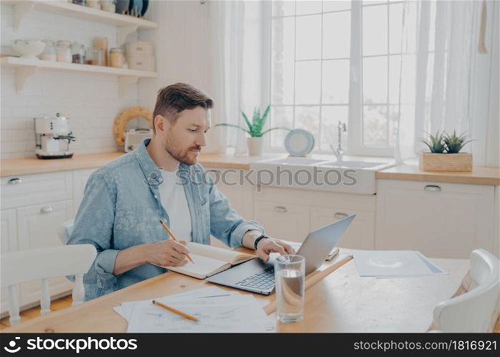 Serious man working on laptop online, sitting at kitchen table and looking at computer screen, focused male writing notes while studying online or searching information for his reports. Serious man working on laptop online at kitchen table