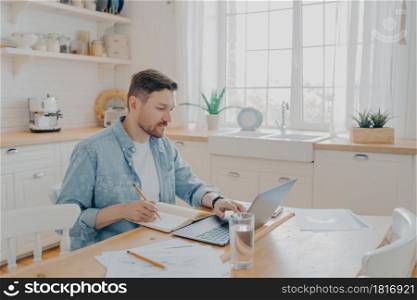 Serious man working on laptop online, sitting at kitchen table and looking at computer screen, focused male writing notes while studying online or searching information for his reports. Serious man working on laptop online at kitchen table