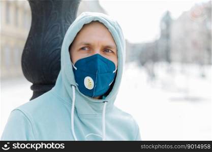 Serious pensive man wears hoodie and protective respiratory mask while walk on street, protects against infection with influenza virus or coronavirus, avoids public crowded places, cares about health