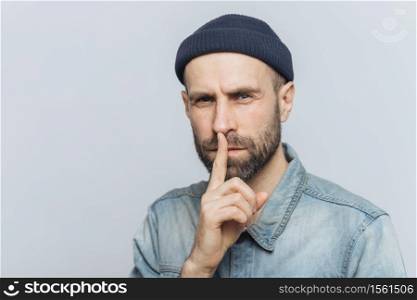 Serious pleasant looking bearded male with mysterious expression keeps fore finger on lips, asks to keep personal information confidential, wears stylish hat and denim shirt, isolated on grey wall