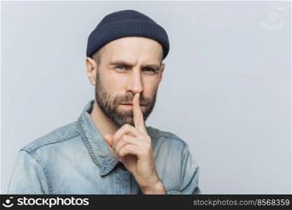 Serious pleasant looking bearded male with mysterious expression keeps fore finger on lips, asks to keep personal information confidential, wears stylish hat and denim shirt, isolated on grey wall