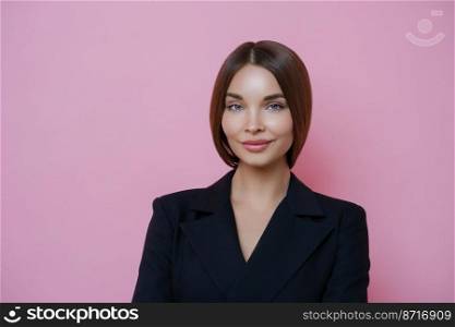 Serious pleased brunette European woman in black clothes stands confident and satisfied, has minimal makeup, appealing look, isolated over pink studio background. Business lady ready for work