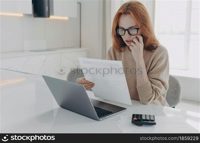 Serious redhead woman focused in paper documents uses calculator and laptop computer for making financial report prepares research poses at desktop against home interior. Family budget and finances. Serious redhead woman focused in paper documents uses calculator and laptop computer