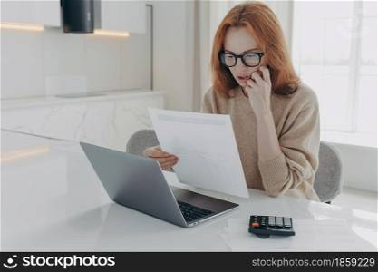 Serious redhead woman focused in paper documents uses calculator and laptop computer for making financial report prepares research poses at desktop against home interior. Family budget and finances. Serious redhead woman focused in paper documents uses calculator and laptop computer
