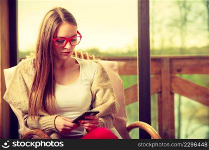 Serious sad woman sitting at home on rocker chair in front of window in her living room using phone. Woman sitting on chair using phone at home