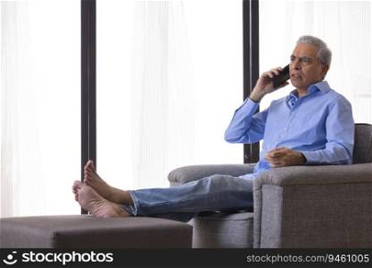 Serious senior man talking on mobile phone while sitting on sofa at home