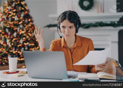 Serious spanish woman teacher in wireless headset with microphone giving online lesson on laptop while working at home office on Christmas, sitting in room with decorated xmas tree on background. Serious spanish woman teacher in headset giving online lesson while working at home on Christmas