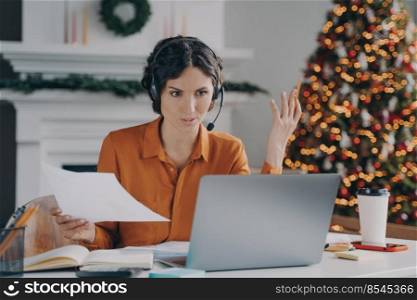 Serious spanish woman teacher in wireless headset with microphone giving online lesson on laptop while working at home office on Christmas, sitting in room with decorated xmas tree on background. Serious spanish woman teacher in headset giving online lesson while working at home on Christmas