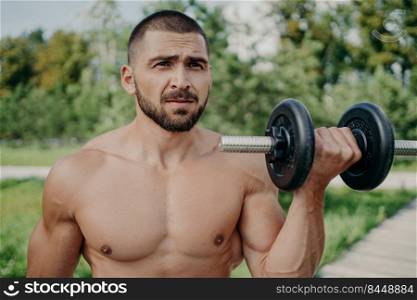 Serious sportsman poses with naked torso has muscular body raises dumbbell, poses outdoor, being in good physical shape, enjoys fresh air and nature. Strength, sporty lifestyle and bodybuilding