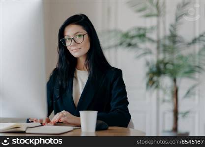 Serious successful female office worker in formal outfit, wears eyeglasses, busy with online work, poses at desktop, works on computer, reads business new or watches tutorial, enjoys working process.