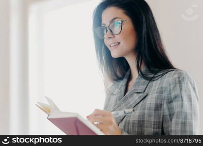 Serious thoughtful brunette European woman writes down text information in notepad, concentrated somewhere into distance, makes notes, wears formal clothes, transparent glasses, stands indoor