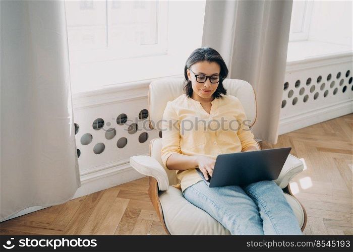 Serious woman freelance employee working at laptop sitting in armchair at home office. Female student wearing glasses using online app for studying. Elearning, remote education, distant job concept.. Female freelance employee working at laptop sitting in armchair at home office. Distant job concept