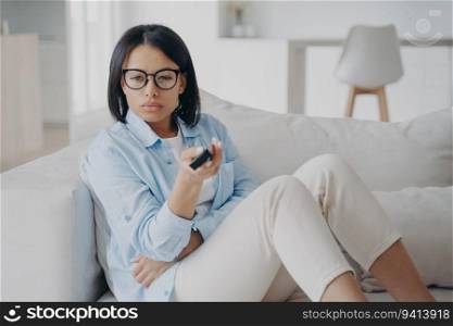 Serious woman in glasses watches TV, changes channels with remote control on couch. Follows live news broadcast.