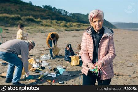 Serious woman showing handful of straws collected on the beach with group of volunteers working in the background. Selective focus in woman in foreground. Woman showing handful of straws collected on the beach