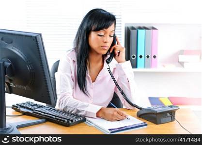 Serious young black business woman on phone taking notes in office