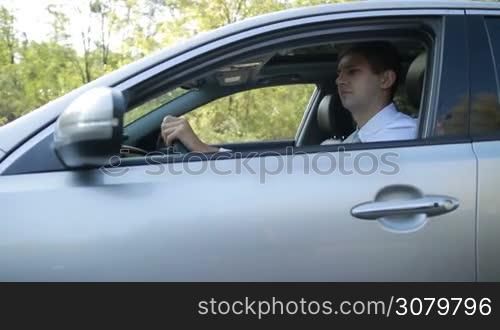 Serious young businessman driving car on rural road in summer during business trip. Confident man enjoying travel on countryside in modern vehicle. Side view. Slow motion. Steadicam stabilized shot.