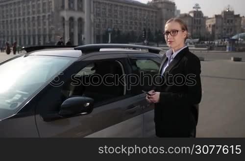 Serious young businesswoman in stylish eyeglasses and formal wear opening car door and getting into automobile. Business woman adjusting car rearview mirror before driving. Slow motion